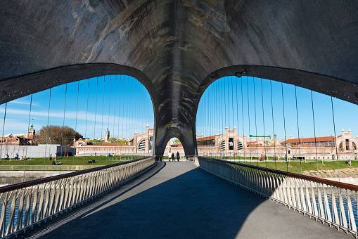 A modern bridge over the Manzanares River in the public park of Madrid Río in Madrid, Spain, with the Matadero building (a former slaughterhouse converted to an arts centre) in the background.